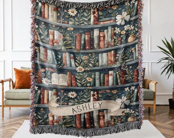 May include: A woven throw blanket featuring a bookshelf design with books, flowers, and the name "ASHLEY" on a banner. The blanket has a dark blue background with fringed edges and is displayed indoors.