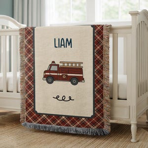 May include: A beige baby blanket with a red plaid border and fringe. The blanket features a maroon fire truck illustration and the name "LIAM" in navy blue. The blanket is draped over a white crib.