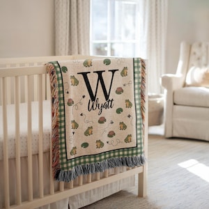 May include: A beige baby blanket with a green and white checkered border and fringe. The blanket features a large black "W" and the name "Wyatt" in black script, along with illustrations of frogs, lily pads, and flowers.
