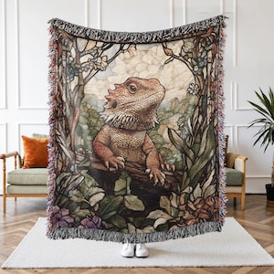May include: A woven throw blanket featuring a detailed illustration of a bearded dragon. The artwork showcases the reptile in a naturalistic setting with lush foliage, in shades of green, brown, and red. The blanket has a fringed edge.