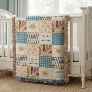 May include: A patchwork baby blanket with a repeating pattern of suns, rainbows, and the text "You Are My Sunshine." The blanket features a fringe edge and is draped over a white crib. The color palette includes beige, blue, orange, and green.