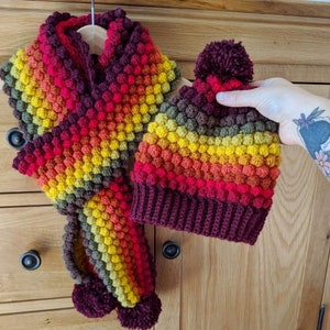 May include: A colourful crocheted hat and scarf set with a bobble on the top of the hat. The set is made with a gradient of red, orange, yellow, and green yarn.
