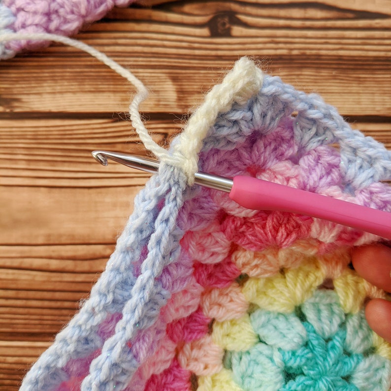 May include: A close-up of a crochet project in progress. The crocheter is working on a corner of a colorful rainbow granny square blanket. The yarn is a light blue color and the crochet hook is pink.