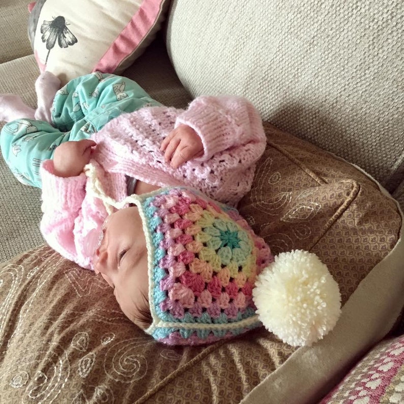 May include: A baby wearing a pink crocheted cardigan and a rainbow crochet hat with a white pom-pom. The baby is lying on a brown patterned couch.