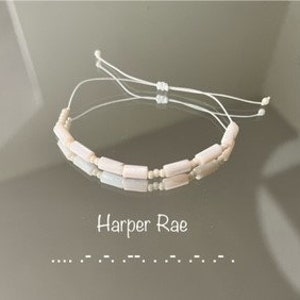May include: A white beaded bracelet with a sliding knot closure. The beads are rectangular and have a subtle shimmer. The bracelet is made with a white cord.
