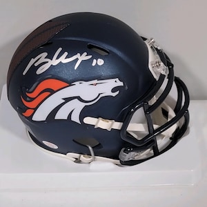 May include: A navy blue football helmet with a white and orange horse logo. The helmet has a black facemask and a signature in white. The helmet is sitting on a white surface.