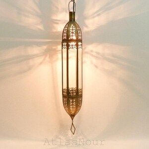 Peut inclure: Une lanterne suspendue en laiton, de forme ovale allongée. Elle présente des motifs ajourés en métal et des panneaux de verre transparents. Un petit pendentif en forme de losange est suspendu en bas. Le texte "AtlasNour" est visible.