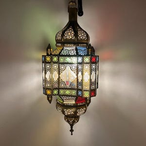 Może przedstawiać: Ozdobna mosiężna lampa wisząca z wielokolorowym witrażem. Lampa posiada skomplikowane wzory wycięć i kopulasty wierzch. Szklane panele są w odcieniach czerwieni, niebieskiego, zieleni i żółci. Ozdobne oświetlenie.