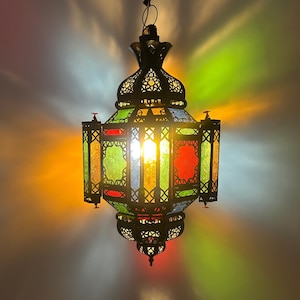 Może przedstawiać: Wisząca lampa w marokańskim stylu z metalową ramą i kolorowymi witrażami. Lampa ma ozdobny szczyt w kształcie ananasa i dekoracyjny wzór.