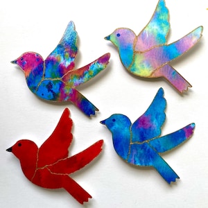 Könnte beinhalten: Vier farbenfrohe Vogel-Ornamente mit goldenen Akzenten. Die Vögel sind in Rot-, Blau-, Rosa- und Lilatönen bemalt.