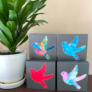 Puede incluir: Cuatro pequeños bloques de madera con un fondo gris oscuro. Cada bloque presenta una silueta de pájaro de color diferente con efectos de pintura tipo acuarela. Los pájaros son en tonos de azul, rosa, rojo y una mezcla de azul, verde y naranja.