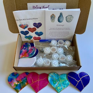 May include: A Kintsugi Heart craft kit in a cardboard box containing instruction booklets, small plastic containers of glue and gloss, a paintbrush, and several heart-shaped cutouts. Four finished heart pieces with gold-colored cracks are displayed in front of the box.