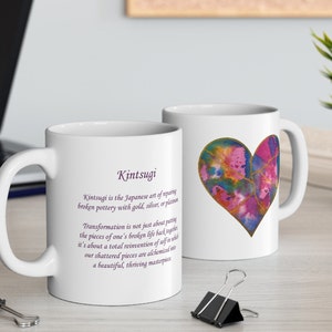 May include: White ceramic mug with a purple heart design and gold accents. The mug features the Japanese word "Kintsugi" and a definition of the art of repairing broken pottery with gold, silver, or platinum.