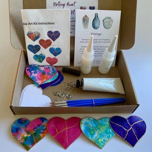 May include: A Kintsugi heart art kit in a cardboard box containing instruction sheets, two white glue bottles, a white paint tube, a black tool, silver wire, blue paintbrushes, and several heart-shaped cutouts with gold-colored crack patterns.