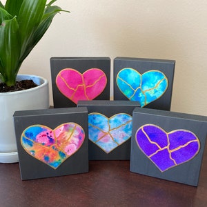 May include: Five small wooden blocks, each painted black and featuring a different watercolor heart design with gold accents. The hearts are pink, blue, purple, and two shades of blue.