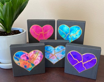 May include: Five small wooden blocks, each painted black and featuring a different watercolour heart design with gold accents. The hearts are pink, blue, purple, and two shades of blue.