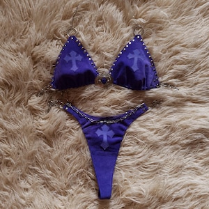 Puede incluir: Conjunto de bikini morado de dos piezas con detalles de cadena plateada. El top triangular y la braguita de estilo tanga presentan adornos en forma de cruz. El bikini está hecho de un material suave similar al ante y adornado con tachuelas plateadas.