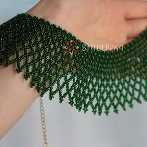 K&ouml;nnte beinhalten: Eine gr&uuml;ne Perlenkette mit einer zarten Kette und einem Herzanh&auml;nger. Die Kette besteht aus kleinen, runden Perlen, die in einem komplizierten Muster miteinander verwoben sind.