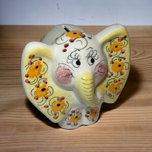 Op de afbeelding: Een witte keramische spaarpot in de vorm van een olifant met gele en oranje bloemmotieven en een roze blos op de wangen.