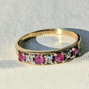 May include: A gold band ring with alternating red and clear gemstones. The ring is set against a textured white surface, with shadows adding depth. The gemstones are round.