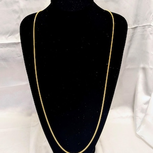 May include: A long, gold-coloured chain necklace displayed on a black mannequin. The chain has a delicate, textured design and hangs vertically, showcasing its length and style. The background is a white fabric.