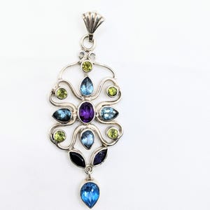 May include: A silver pendant with an elaborate design, set with a variety of colourful gemstones. The pendant features a large, teardrop-shaped blue stone at the base, with other blue, green, and purple stones throughout the design.