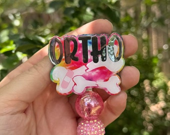 Könnte beinhalten: Ein rosa-weißer Batik-Anhänger mit dem Wort "ORTHO" und einem gebrochenen Knochen mit einem Verband. Der Anhänger ist an einem rosa-weißen Perlen-Schlüsselanhänger befestigt.