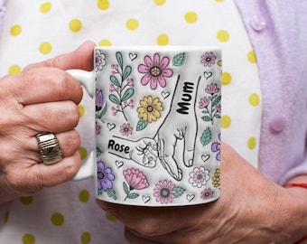 May include: A white ceramic mug featuring a colorful floral pattern with pink, yellow, purple, and teal flowers. The design includes a line art illustration of an adult hand holding a child's hand, with the text Mum and Rose printed on them.