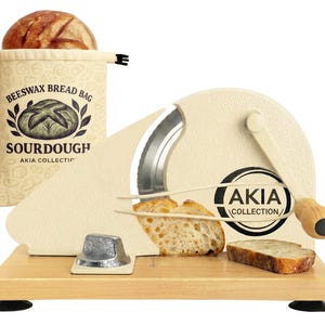 May include: A cream-colored bread slicer with a wooden base and handle, featuring a curved blade. A loaf of bread sits in a beige beeswax bread bag labeled "Sourdough." The slicer has the "AKIA COLLECTION" logo.