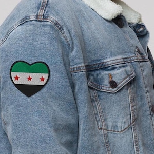 Peut inclure: Veste en jean bleu clair avec un col en fausse fourrure blanche. Un écusson en forme de cœur avec le drapeau syrien est cousu sur le côté gauche. La veste a une fermeture boutonnée et deux poches de poitrine.