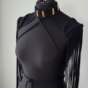 May include: A black macrame harness featuring a high neck collar with gold-toned cylindrical accents, braided straps crossing the chest, and long fringe detailing on the shoulders. The piece is displayed on a black mannequin torso with white pins.