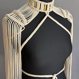 May include: A cream-colored macrame body harness featuring a high-neck choker collar with integrated gold-toned metal accents. The design includes braided cord straps that cross the chest and waist, with long, flowing fringe extending over the shoulders.