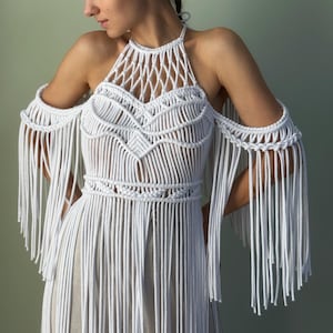 Puede incluir: Un top halter de macramé blanco con intrincados patrones de nudos en el pecho y la cintura, con largos flecos fluidos que se extienden desde los hombros por los brazos y el torso, creando una apariencia texturizada y superpuesta.