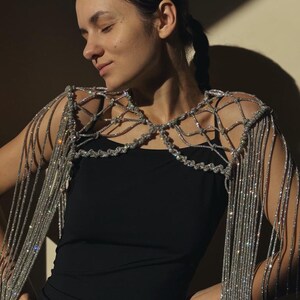 May include: A silver crystal-embellished shoulder accessory featuring a lattice-style net design across the chest and shoulders, with long, cascading silver fringe strands hanging from the edges, worn over a simple black sleeveless garment.