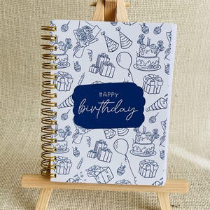 May include: A white spiral-bound notebook with a birthday pattern of cakes, gifts, and balloons. The words "HAPPY birthday" are written in a dark blue banner. The notebook is displayed on a small wooden easel.