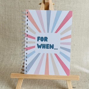May include: A notebook with a spiral binding, featuring a vibrant sunburst pattern in shades of pink, peach, blue, and beige. The cover displays the text "FOR WHEN...". It's presented on a small wooden easel, ideal for a desk or table.