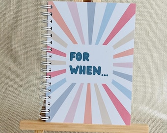May include: A spiral-bound notebook featuring a sunburst pattern in shades of pink, blue, and beige. The centre of the cover displays the text "FOR WHEN..." in bold, dark blue letters. The notebook is displayed on a small wooden easel.