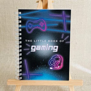 May include: A spiral-bound notebook with a black cover featuring neon blue and pink graphics of a game controller and a headset. The text on the cover reads THE LITTLE BOOK OF gaming in white and stylised lettering against a dark background.