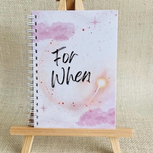 May include: A spiral-bound notebook featuring a white cover with pink clouds, a glowing celestial arc, and small stars. The text "For When" is printed in black script in the centre. The notebook is displayed on a small wooden easel.