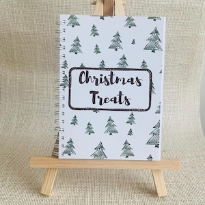 May include: A spiral-bound notebook with a white cover featuring a repeating pattern of dark green hand-drawn pine trees. The centre of the cover displays a dark brown rectangular frame containing the text Christmas Treats.