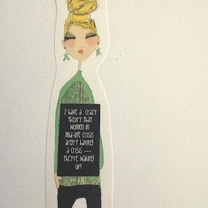 May include: A die-cut bookmark featuring an illustration of a person with a high yellow bun hairstyle, green earrings, a green sweater, and black pants. A black rectangular sign displays the text: I have a crazy theory that women in mid-life crisis aren't having a crisis --- they're waking up!