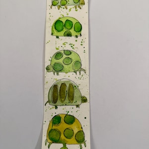 May include: A handmade bookmark featuring watercolor illustrations of green turtles. Each turtle has a unique shell pattern and is painted in shades of green and yellow. The bookmark is decorated with green paint splatters.