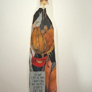 May include: A tall, narrow bookmark featuring an illustration of a person with a high bun hairstyle, wearing a black garment. Below, large orange and brown floral shapes surround a red rooster. The text reads: The older I get, the more I understand why roosters scream to start their day.
