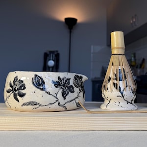 May include: A ceramic matcha set featuring a bowl, a whisk holder, and a bamboo scoop. The bowl and holder are cream-colored with black speckled patterns and hand-painted black floral designs. The bamboo whisk is placed inside the holder.