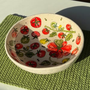 May include: A round, off-white ceramic bowl with a speckled finish, featuring hand-painted illustrations of red tomatoes on green vines with small yellow flowers. The interior of the bowl is decorated with multiple tomato clusters.