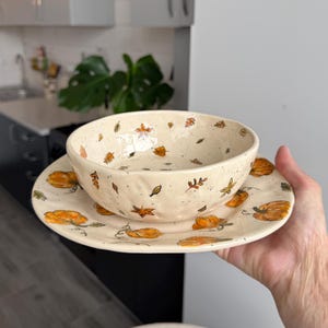 Może przedstawiać: Kremowy zestaw ceramiczny składający się z miski i pasującego talerza, ozdobiony ręcznie malowanym wzorem pomarańczowych dyń i rozrzuconych jesiennych liści. Miska ma teksturowaną, lekko nierówną krawędź, a na talerzu widoczne są większe ilustracje dyń.