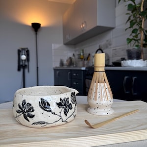 May include: A ceramic matcha bowl with a cream-colored glaze and black speckled finish, featuring hand-painted black floral motifs. Beside it sits a bamboo matcha whisk on a matching speckled stand and a long, curved bamboo tea scoop.