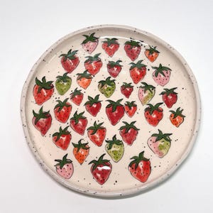Peut inclure: Une assiette ronde en céramique avec une surface mouchetée de couleur crème. L'assiette est décorée d'illustrations à l'aquarelle de fraises de différentes tailles et couleurs, dont le rouge, le rose, le vert et l'orange. Les fraises ont des tiges vertes.
