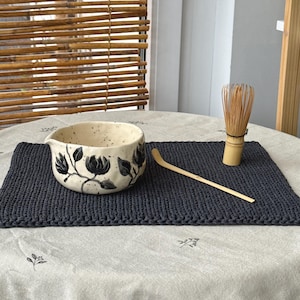 May include: A ceramic matcha bowl with black floral designs, a bamboo whisk, and a wooden scoop rest on a dark gray knitted placemat. The items are arranged on a round table covered with a light gray tablecloth.