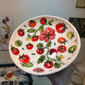 May include: A round ceramic bowl with a cream-coloured base featuring a hand-painted pattern of red tomatoes, green leaves, and small yellow flowers. The surface has a speckled texture with dark brown dots scattered across the interior.